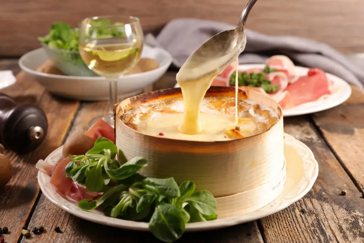 Un plat de fromage fondu, une spécialité du Jura, servi avec des pommes de terre, de la charcuterie et une salade verte. Un verre de vin blanc complète ce festin.