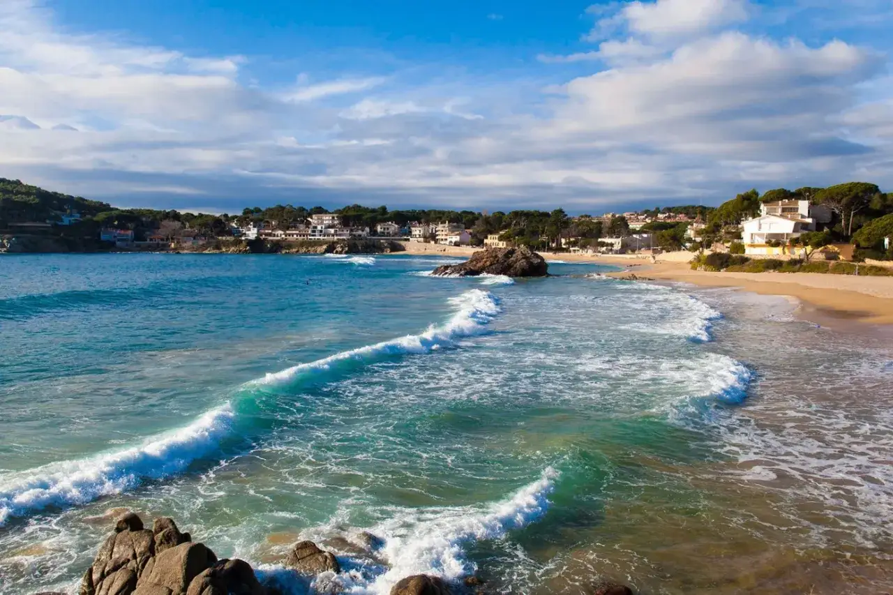 T&uuml;rkisblaue Wellen brechen sich an einem Sandstrand in Palamos, Spanien. Wei&szlig;e H&auml;user und gr&uuml;ne H&uuml;gel s&auml;umen die K&uuml;ste.