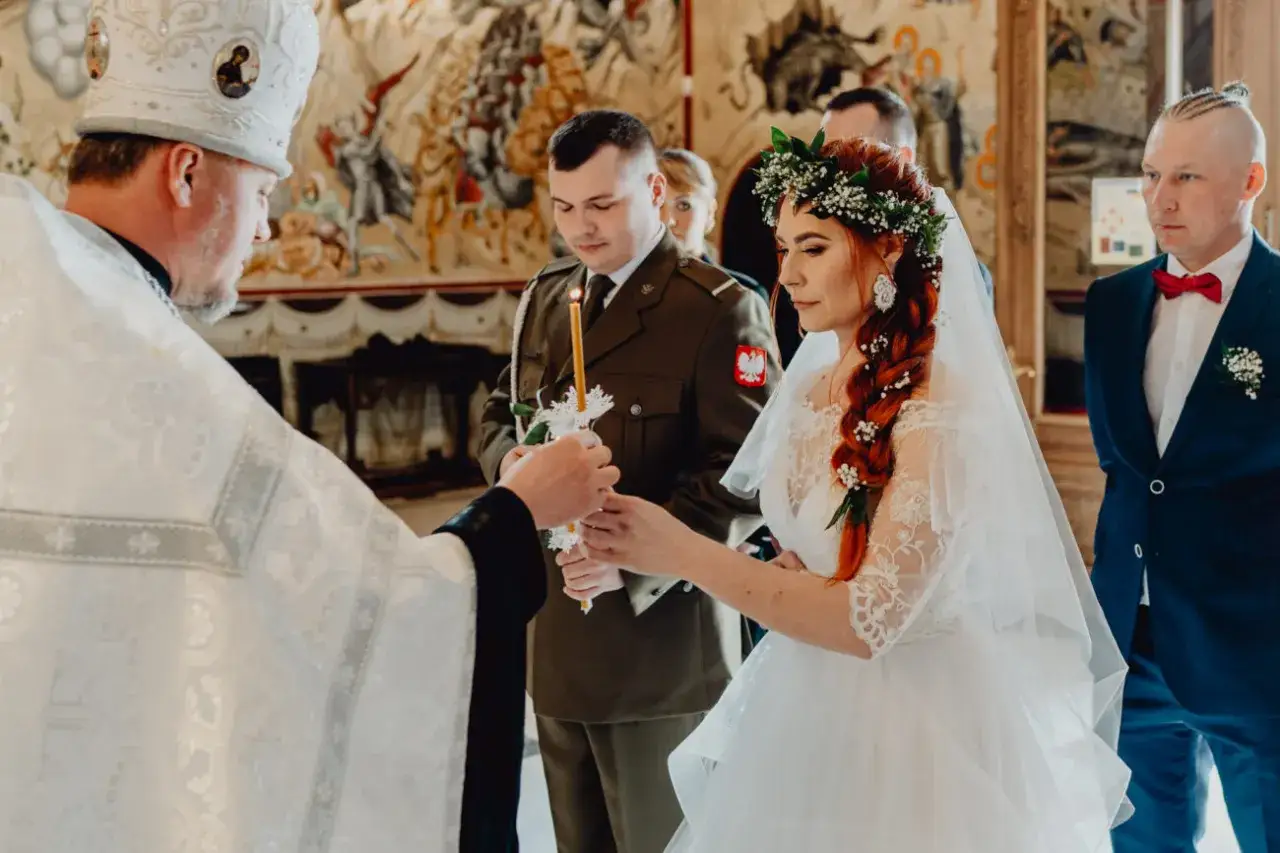 Piękny ślub prawosławny. Panna młoda w wianku, pan młody w mundurze wojskowym. Duchowny błogosławi parę przy świecach.