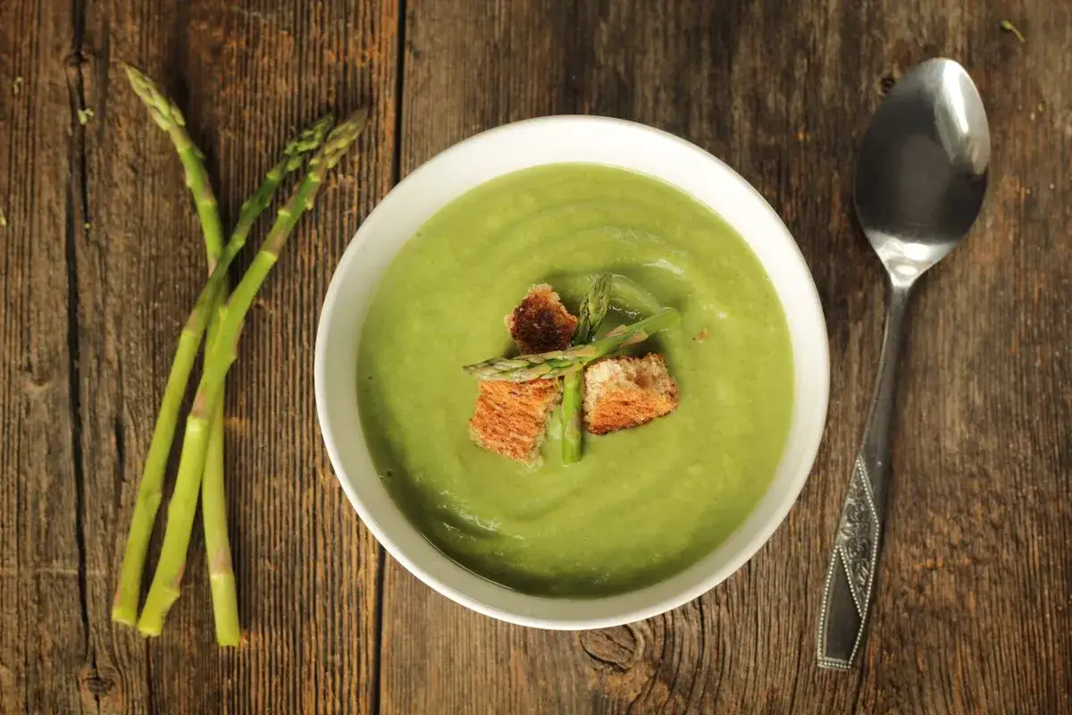 Una ciotola di cremosa crema di asparagi, guarnita con crostini e asparagi freschi, accanto a un mazzo di asparagi su un tavolo di legno rustico.