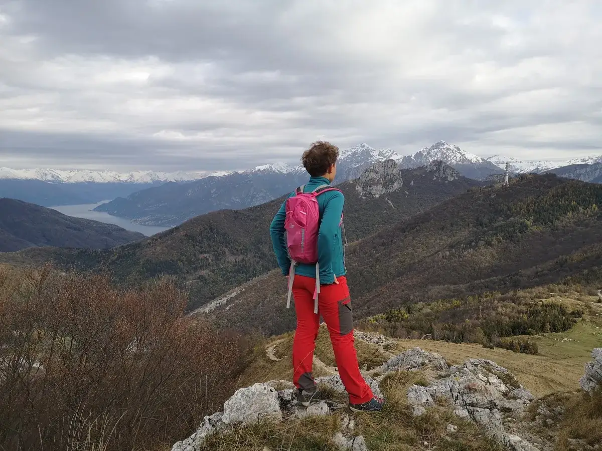 Osoba w czerwonych spodniach i turkusowej bluzie z plecakiem podziwia widok na g&oacute;ry i jezioro. To właśnie jest hiking!