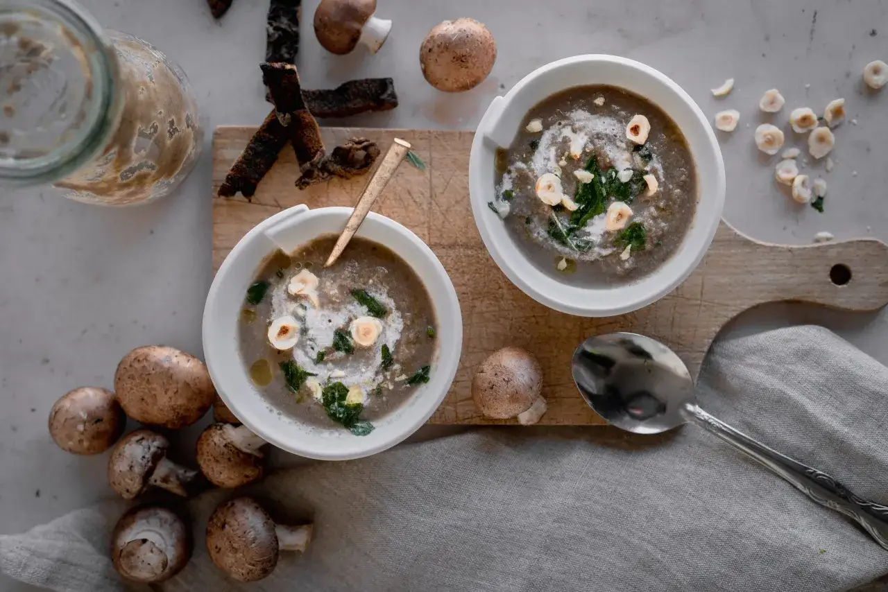 Deux bols de velouté de champignon, garni de crème, de persil et de noisettes. Des champignons frais et des croûtons sont disposés autour.