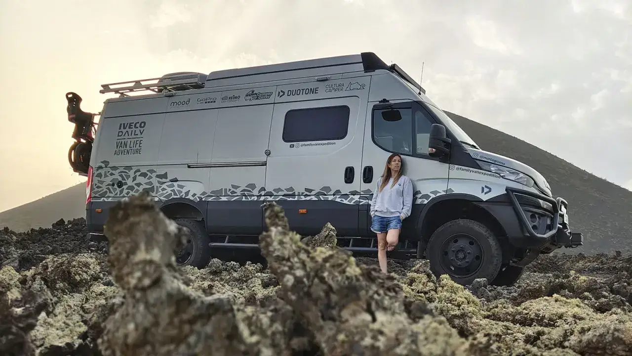Une femme se tient à côté d'un **van tout terrain** IVECO DAILY, prêt pour une aventure en vanlife sur un terrain rocailleux.