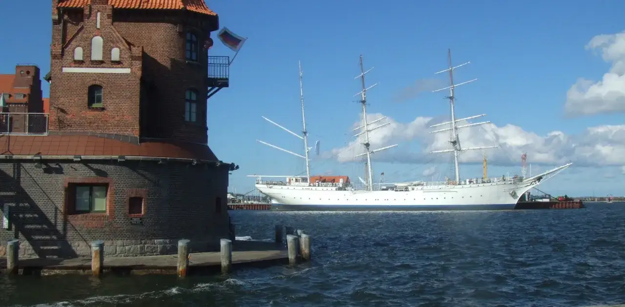Das Segelschulschiff Gorch Fock in Stralsund, ein imposantes weißes Schiff mit drei Masten, liegt vor einem Backsteinturm im Hafen.