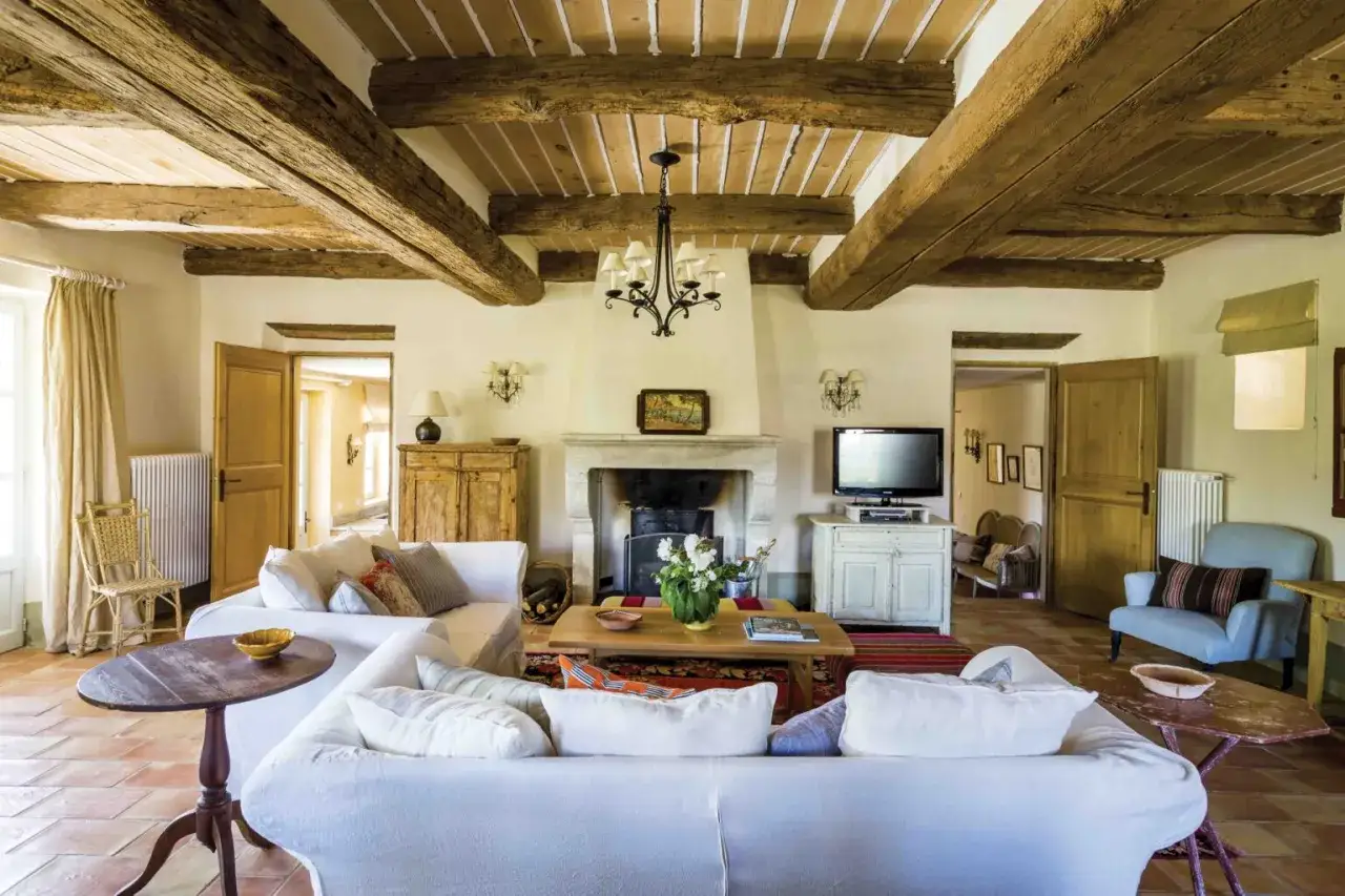 Salon d'une maison provençale avec poutres apparentes, cheminée en pierre, canapés blancs confortables et sol en terre cuite.