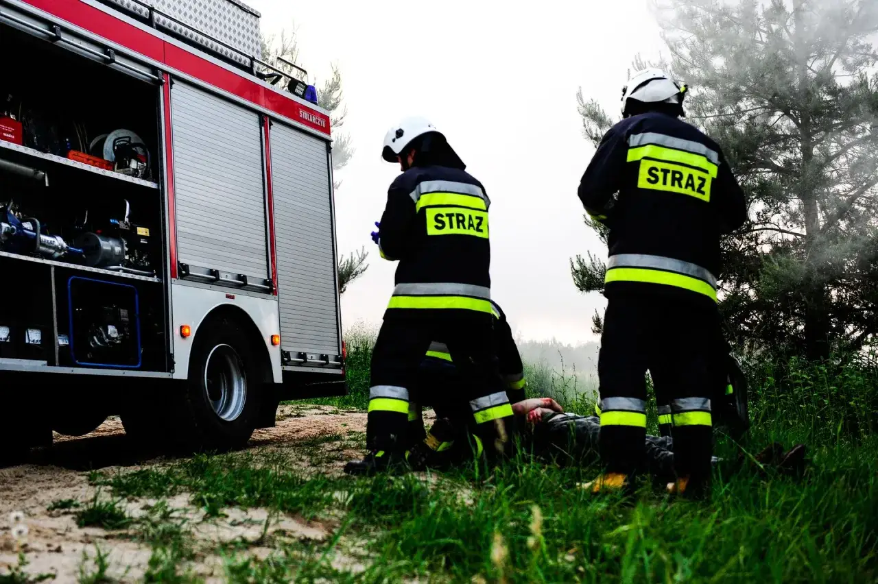 Strażacy przy wozie bojowym udzielają pomocy poszkodowanemu. Sytuacja przypomina trudną akcję, być może ostatnią przed emeryturą strażaka.
