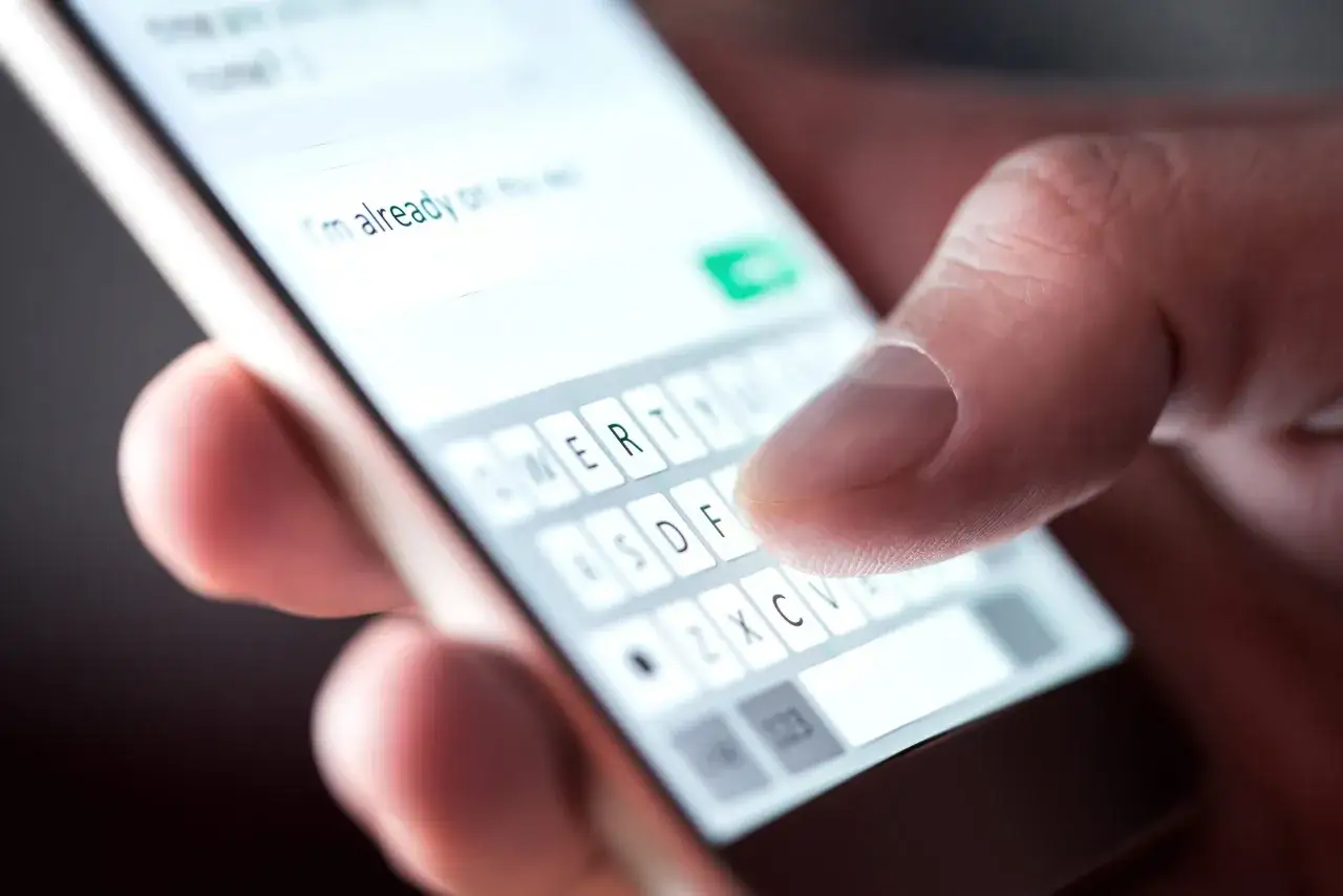 Dłoń pisze wiadomość na telefonie, używając klawiatury ekranowej. Widać fragment tekstu