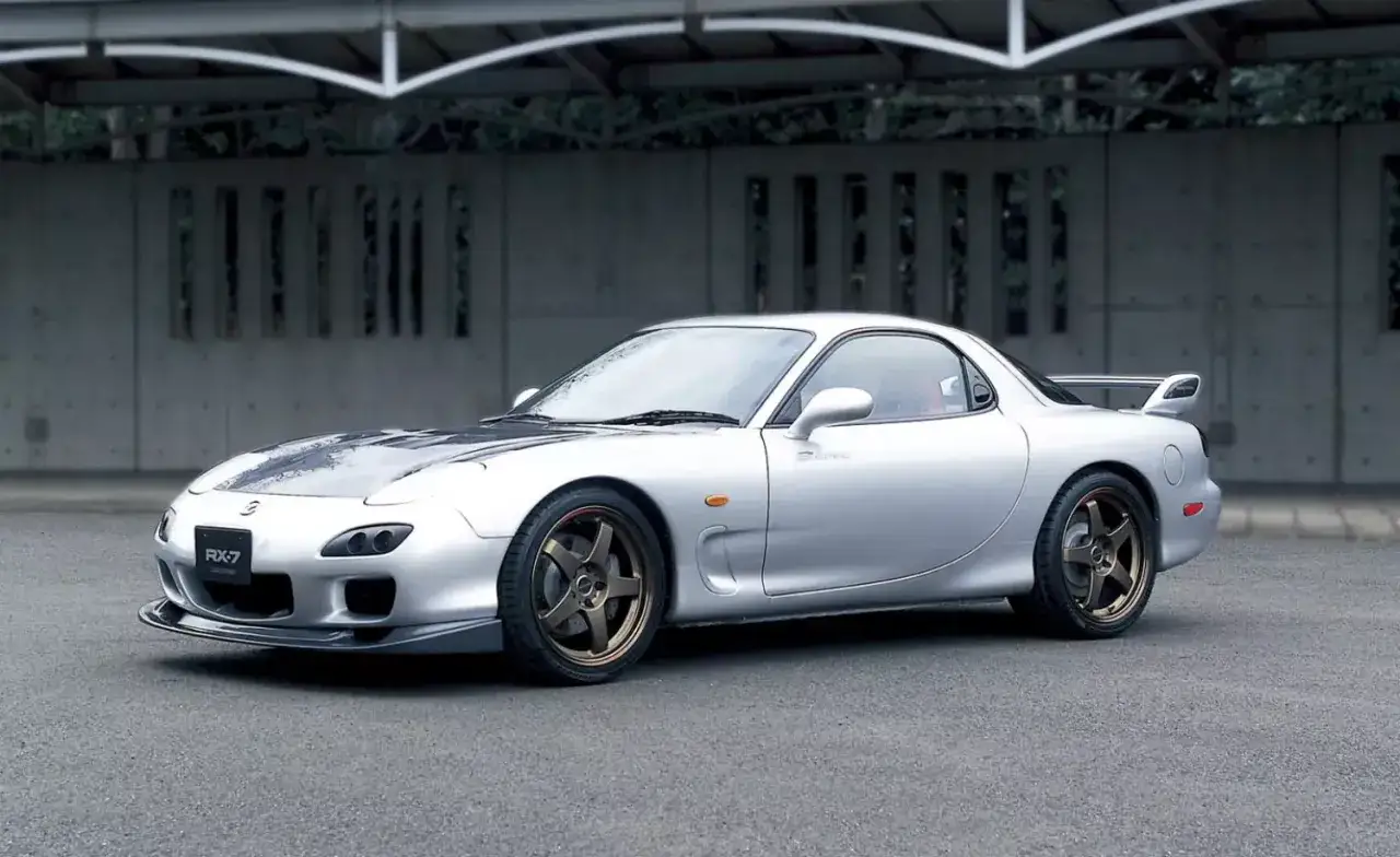 Srebrne auto sportowe z silnikiem Wankla, Mazda RX-7, z przyciemnianą maską i brązowymi felgami, stoi na asfalcie.