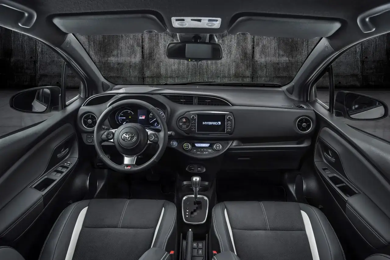 Wnętrze Toyota Yaris GR Sport: sportowa kierownica, ekran multimedialny z napisem HYBRID, czarne fotele z białymi wstawkami.