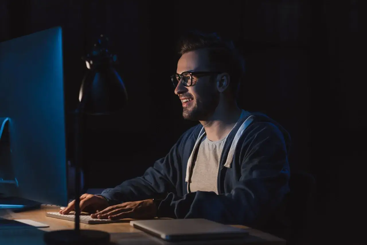 Mężczyzna w okularach z uśmiechem pracuje przy komputerze, ucząc się do studiów podyplomowych tester oprogramowania.