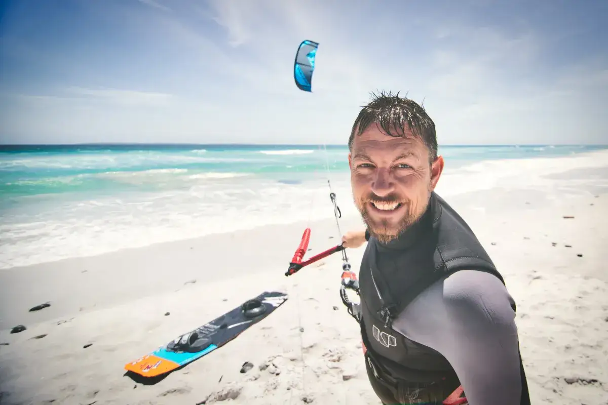 Ein Mann mit einem breiten L&auml;cheln steht am Strand, bereit zum **surfen mit drachen in der hand**. Ein Kiteboard liegt im Sand, ein blauer Kite schwebt am Himmel.