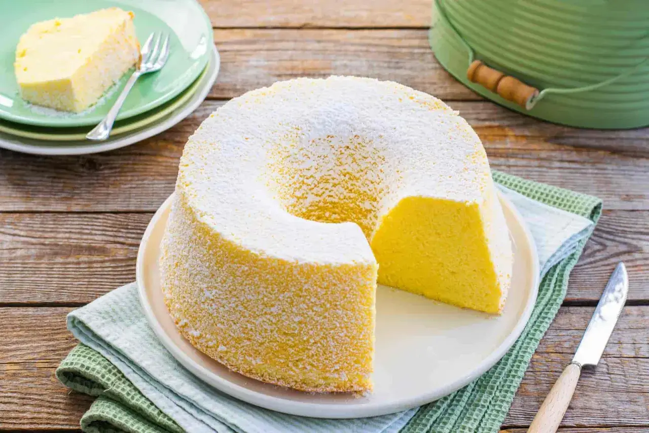 Un soffice chiffon cake, spolverato di zucchero a velo, attende di essere gustato. Una fetta è già su un piattino verde.