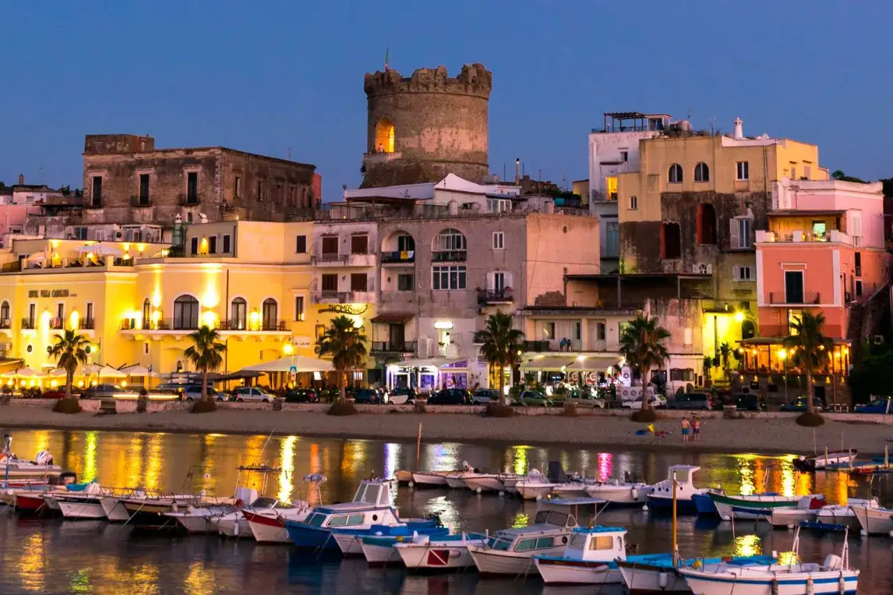 Il torrione di Forio domina il lungomare illuminato, con barche ormeggiate nell'acqua scintillante.