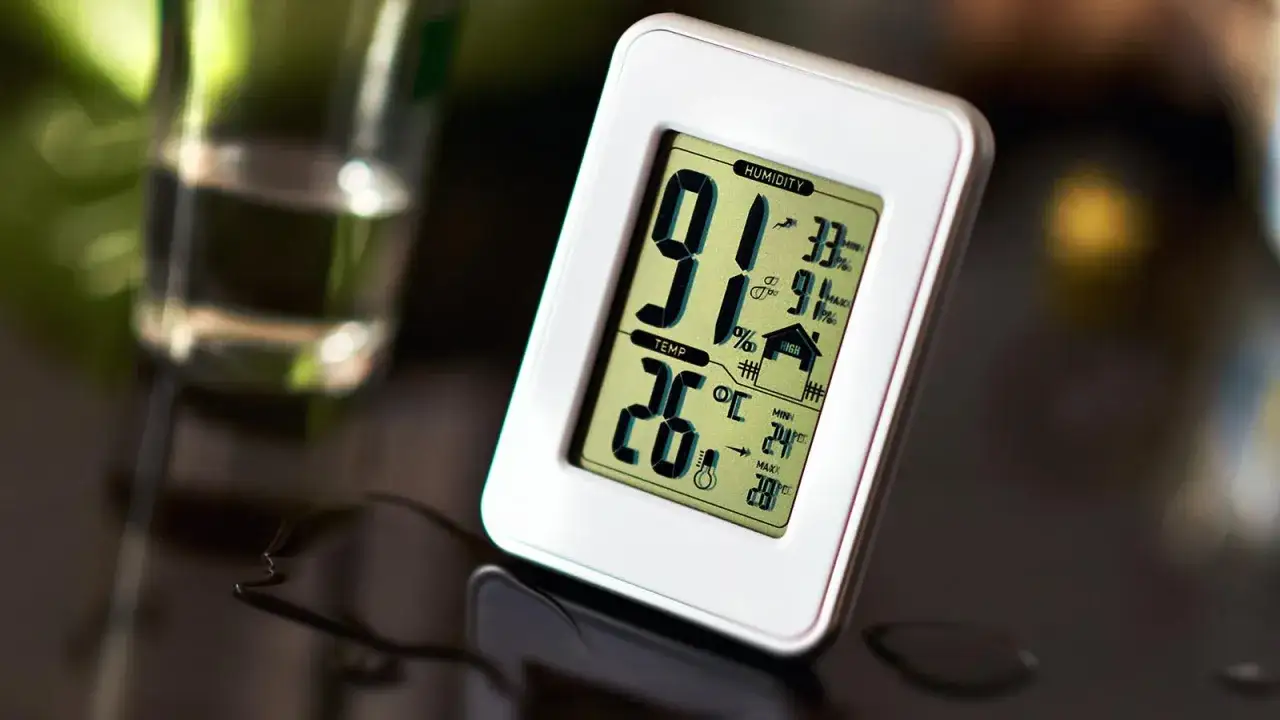 Un higrómetro digital muestra 91% de humedad y 26°C de temperatura. Un vaso con agua y gotas de humedad completan la escena.