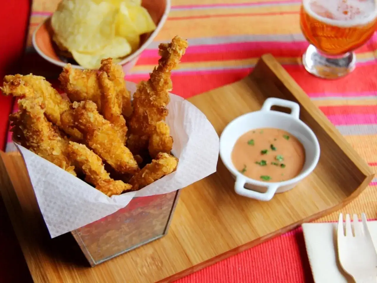 Crujientes tiras de pollo frito servidas en un cubo met&aacute;lico, acompa&ntilde;adas de salsa rosa y patatas fritas.