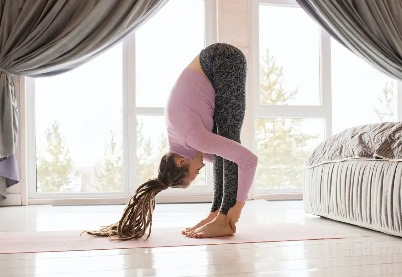 Mujer con rastas en posición de uttanasana yoga, con las manos sujetando los tobillos y la cabeza inclinada hacia el suelo.