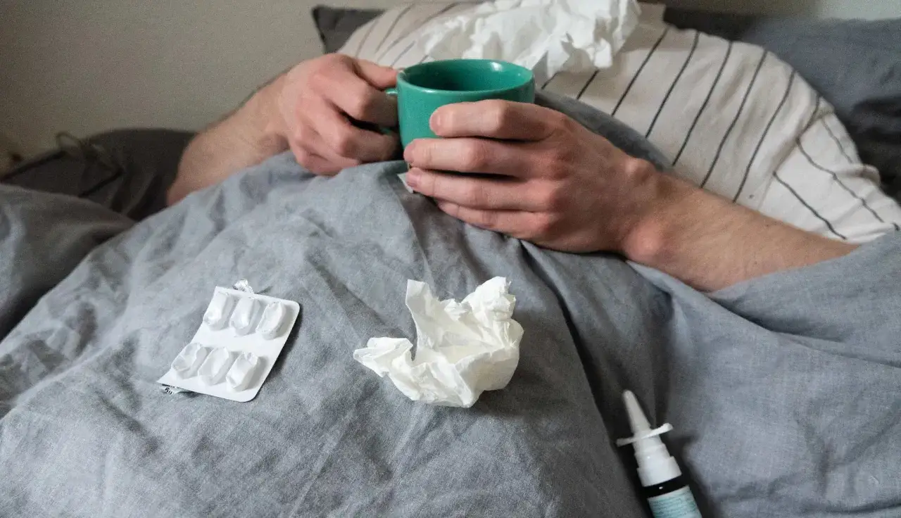 Personne malade au lit, avec une tasse, des mouchoirs, un spray nasal et une boîte de pilules. Des indices sur comment guerir du covid.