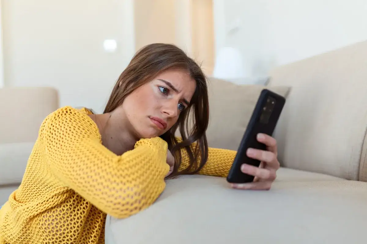 Zamyślona dziewczyna w żółtym swetrze, patrząca na telefon. Czy to znak, jak nadmierny czas przed ekranem wpływa na organizm?