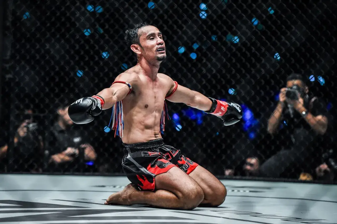 A triumphant strawweight boxer kneels in the ring, arms outstretched, celebrating a hard-fought victory.