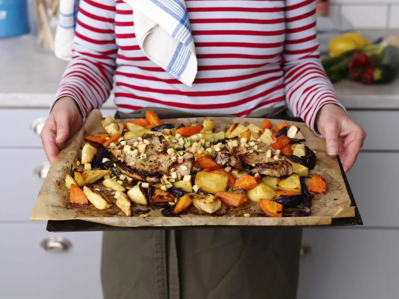 En person h&aring;ller en pl&aring;t med saftiga rotfrukter i ugn, blandat med k&ouml;tt och &auml;pple.