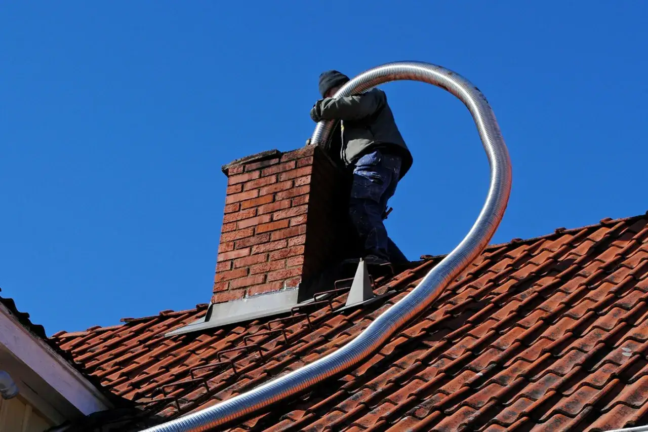 Un couvreur installe un tubage de cheminée sur un toit en tuiles rouges, sous un ciel bleu clair.