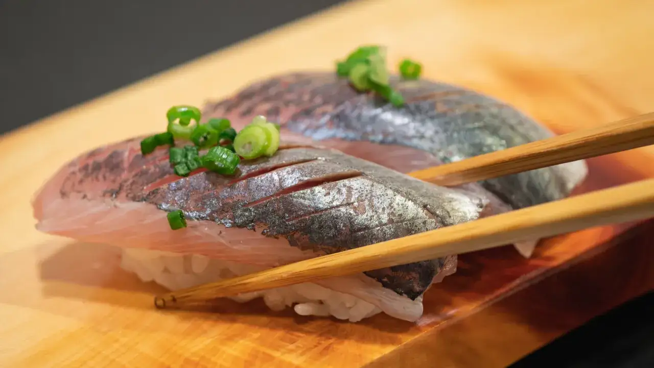 Dwa kawałki sushi z japońskimi rybami, posypane szczypiorkiem, trzymane pałeczkami na drewnianej desce.