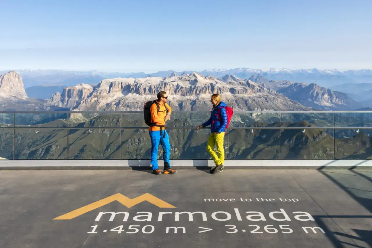 Zwei Wanderer auf einer Aussichtsplattform mit Blick auf die Dolomiten. Der Schriftzug 