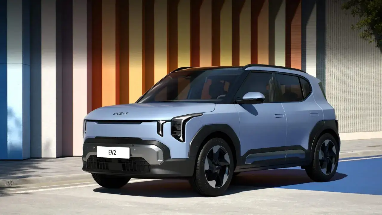 La Kia EV2, un SUV elettrico dal design audace, si presenta in un blu pastello con dettagli grigi e neri, parcheggiata davanti a un muro a strisce colorate.