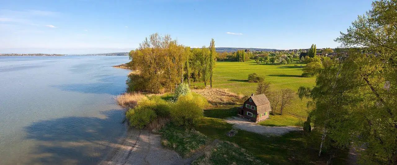 Ein kleines Haus am Ufer des Eriskircher Rieds, umgeben von grünen Wiesen und Bäumen, mit Blick auf den See.