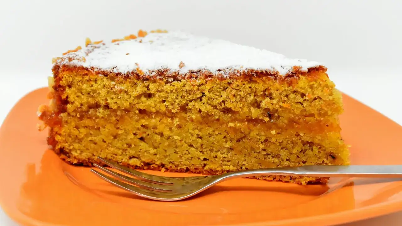 Wilgotne ciasto marchewkowe fit, posypane cukrem pudrem, czeka na widelcu na pomarańczowym talerzu.