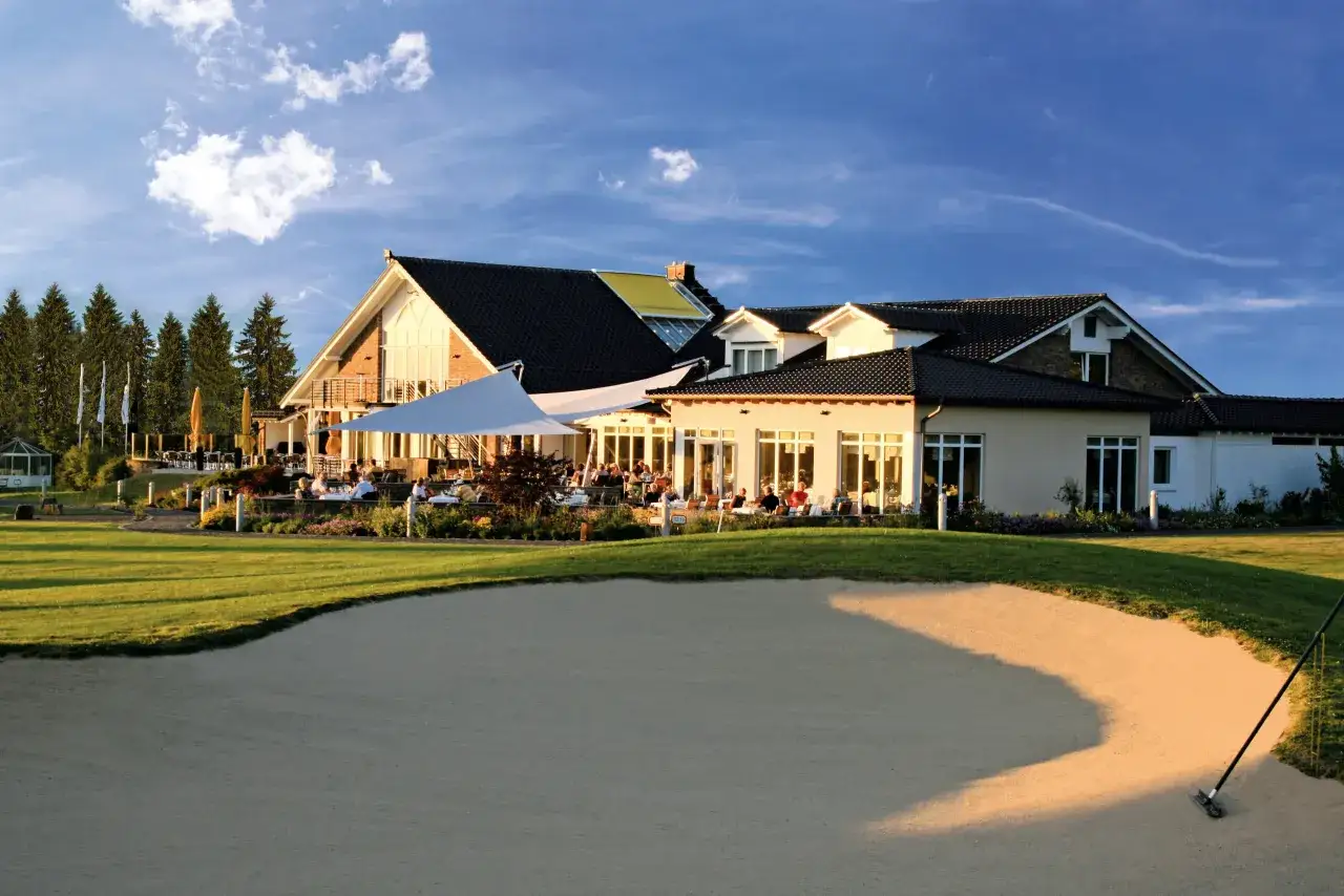 Sonnenuntergang am Golfclub Heckenhof. G&auml;ste genie&szlig;en den Abend auf der Terrasse vor dem Clubhaus, umgeben von gepflegtem Gr&uuml;n und einem Sandbunker.