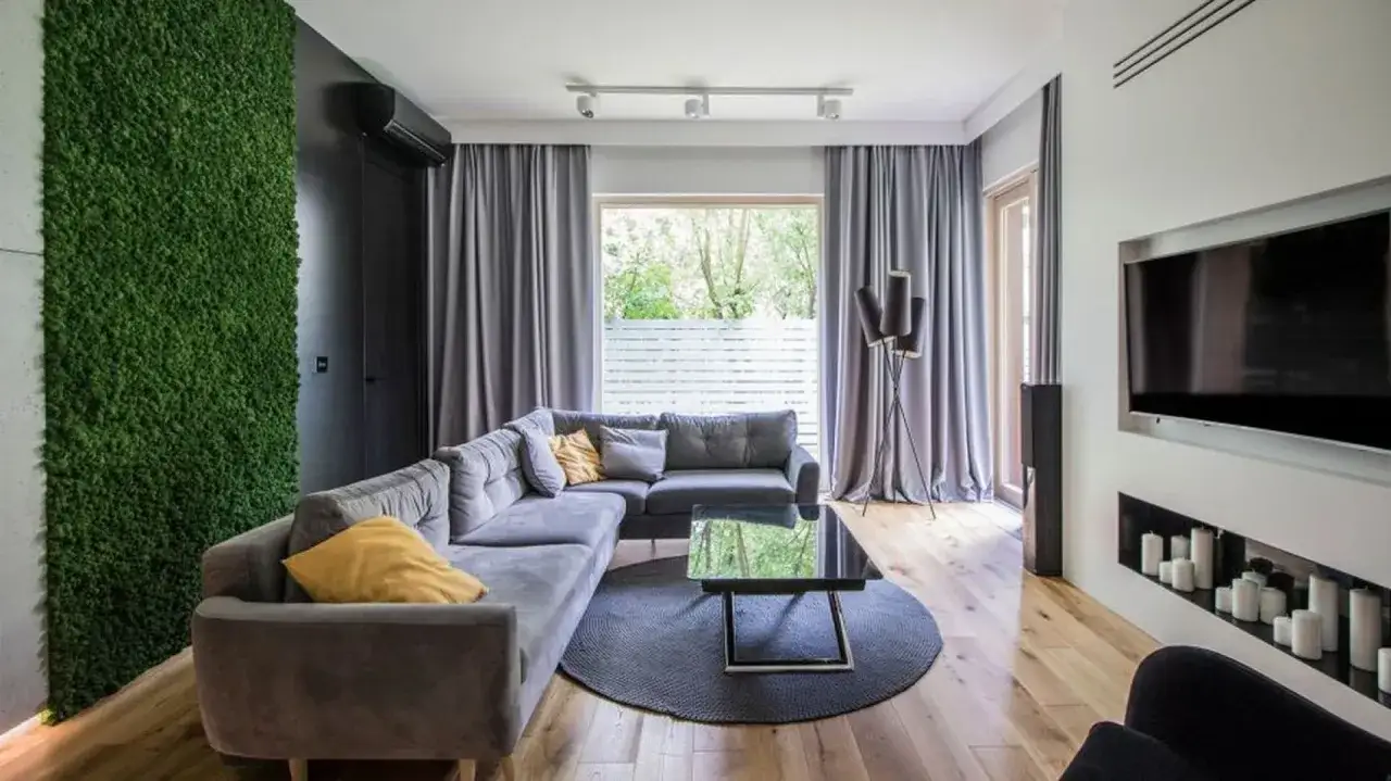 Szary salon zyskuje ciepło dzięki zielonej ścianie z mchu i ż&oacute;łtym poduszkom. Nowoczesny design z szarą sofą i drewnianą podłogą.