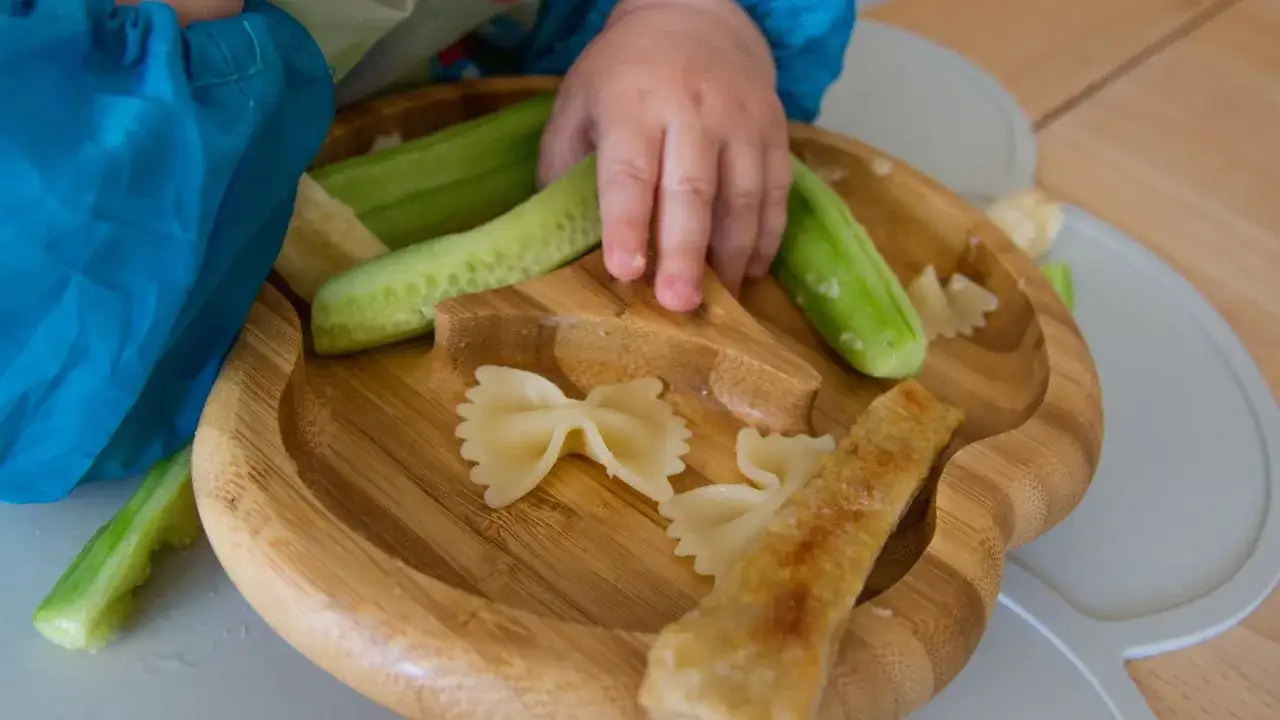 Manina esplora il piatto con bastoncini di cetriolo e pasta, un'idea per ricette svezzamento 6 mesi.