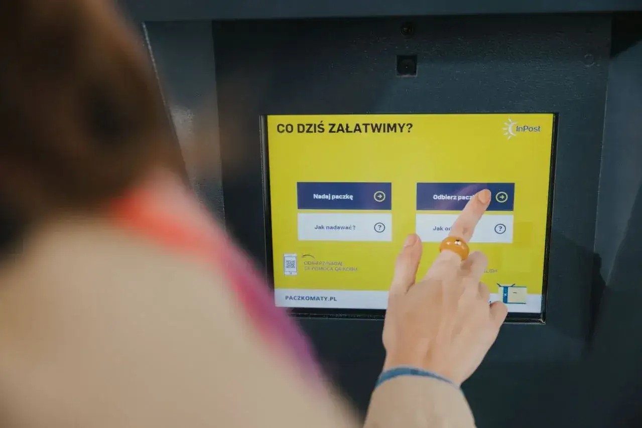 Dłoń z pierścionkiem dotyka ekranu paczkomatu InPost, wybierając opcję odbioru paczki. To szybkie i wygodne rozwiązanie, wspierane przez fundację.