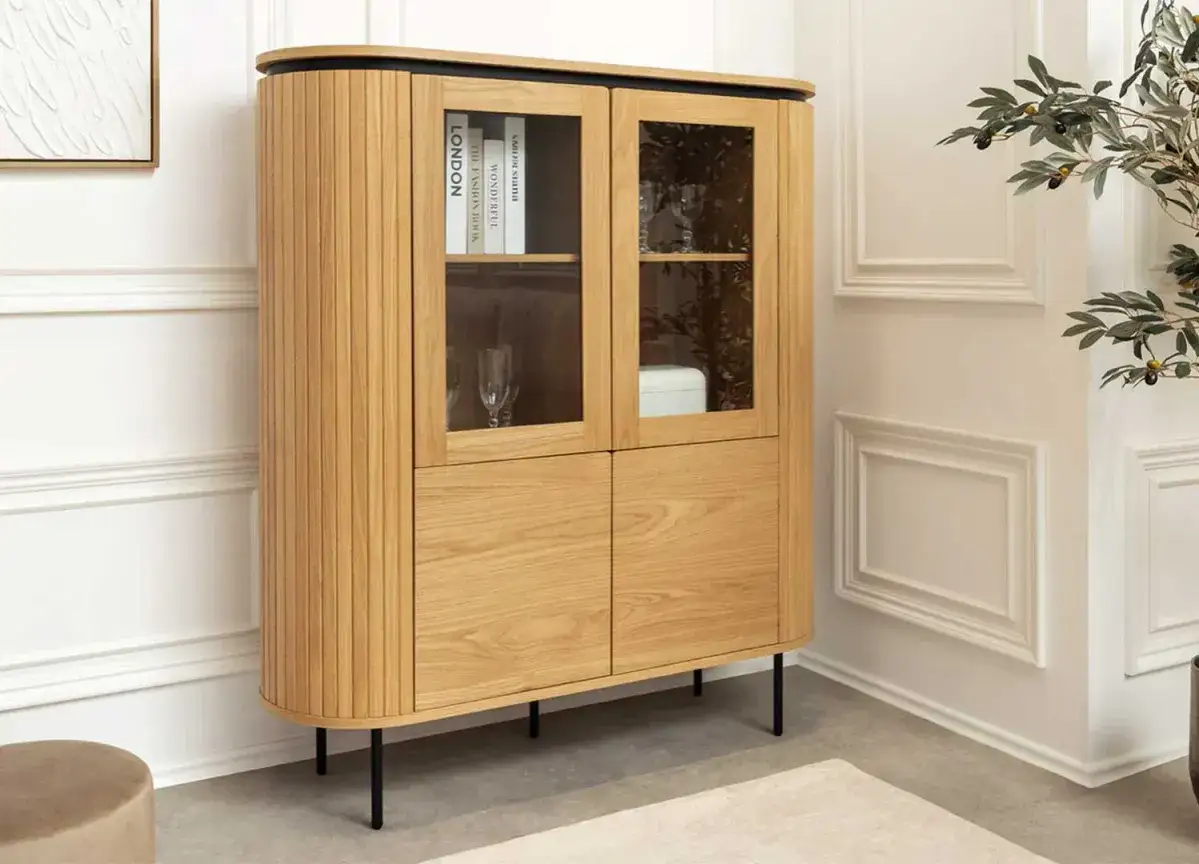 Buffet haut en bois clair avec portes vitrées exposant des livres et des verres. Ses pieds fins noirs ajoutent une touche moderne.