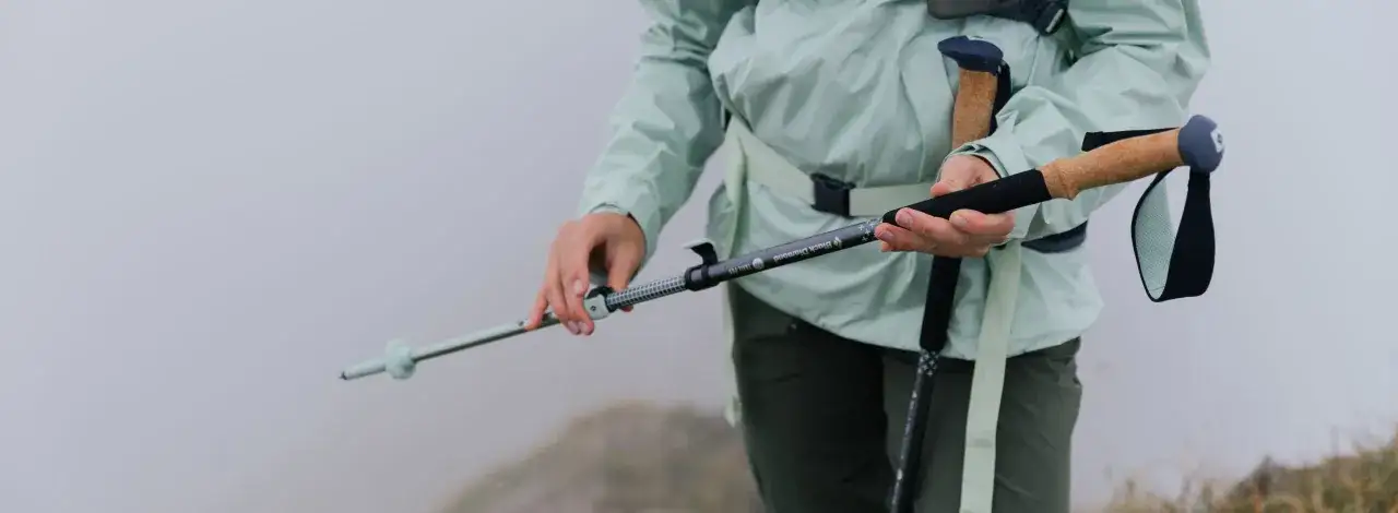 Osoba w jasnej kurtce trzyma kijki trekkingowe Black Diamond Trail, gotowa na wędrówkę po mglistym szlaku.