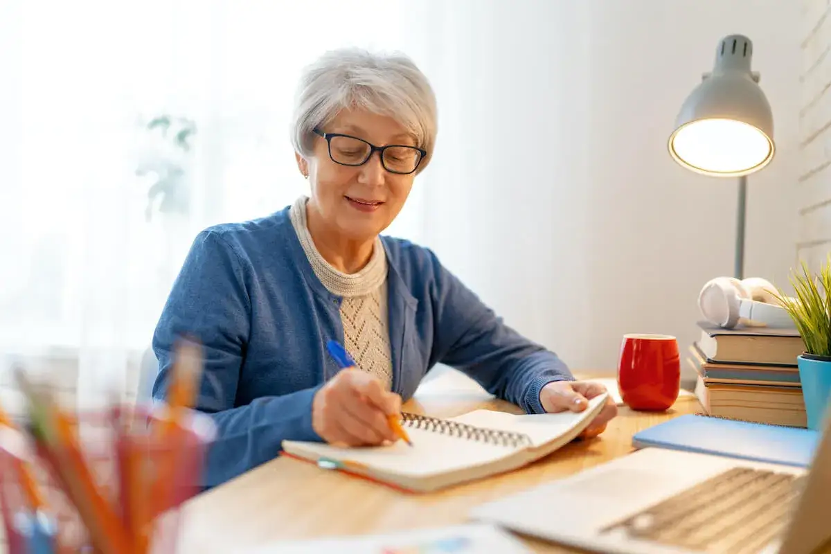 Uśmiechnięta seniorka pisze w notatniku, ciesząc się ulgą dla pracujących seniorów. Na biurku lampka, kubek i książki.