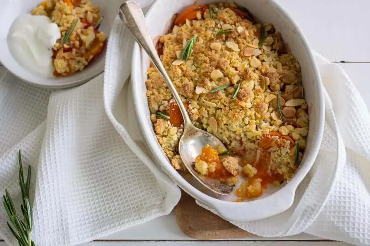 Un d&eacute;licieux crumble abricot aux amandes effil&eacute;es et romarin, servi dans un plat blanc avec une cuill&egrave;re.