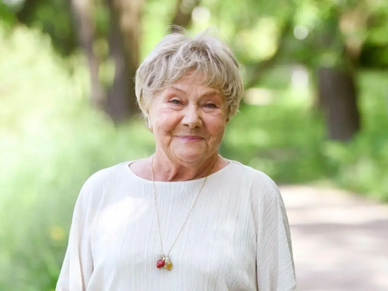 Uśmiechnięta Teresa Lipowska, której wiek budzi podziw, w białej bluzce z naszyjnikiem.