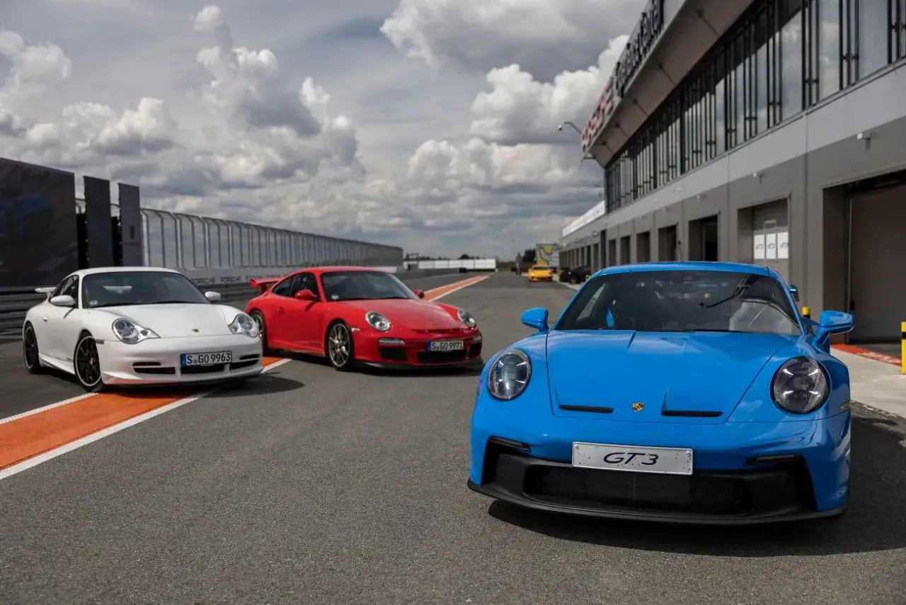 Trzy Porsche 911: białe, czerwone i niebieskie GT3, stoją na torze wyścigowym.