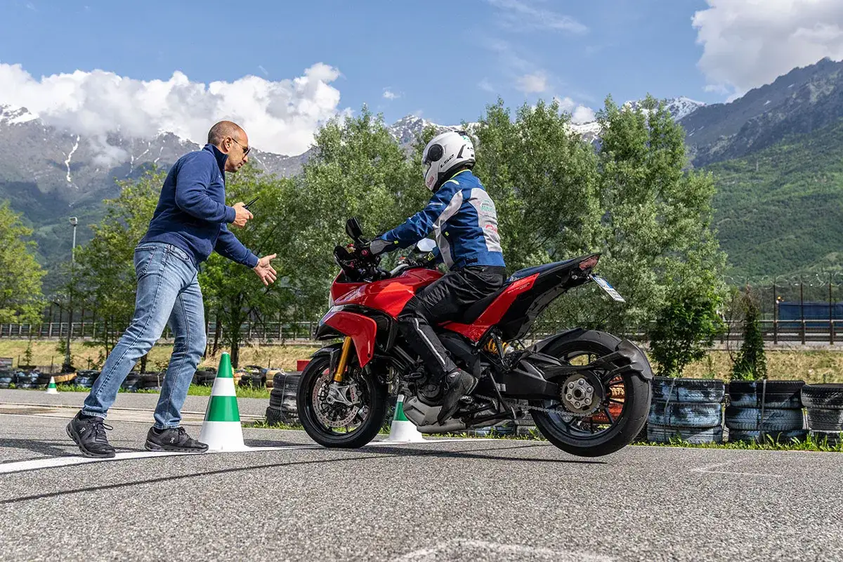 Istruttore spiega come guidare una moto a un allievo su una Ducati rossa, con montagne sullo sfondo.