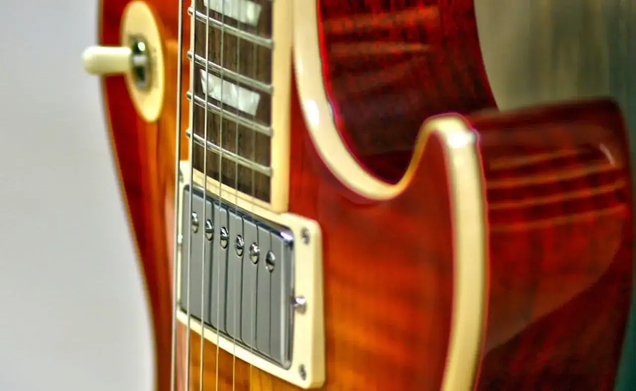 Część korpusu i gryfu gitary elektrycznej w odcieniach czerwieni i brązu. Widoczne przetworniki, struny i przełącznik. Klasyczne marki gitar elektrycznych inspirują muzyków.