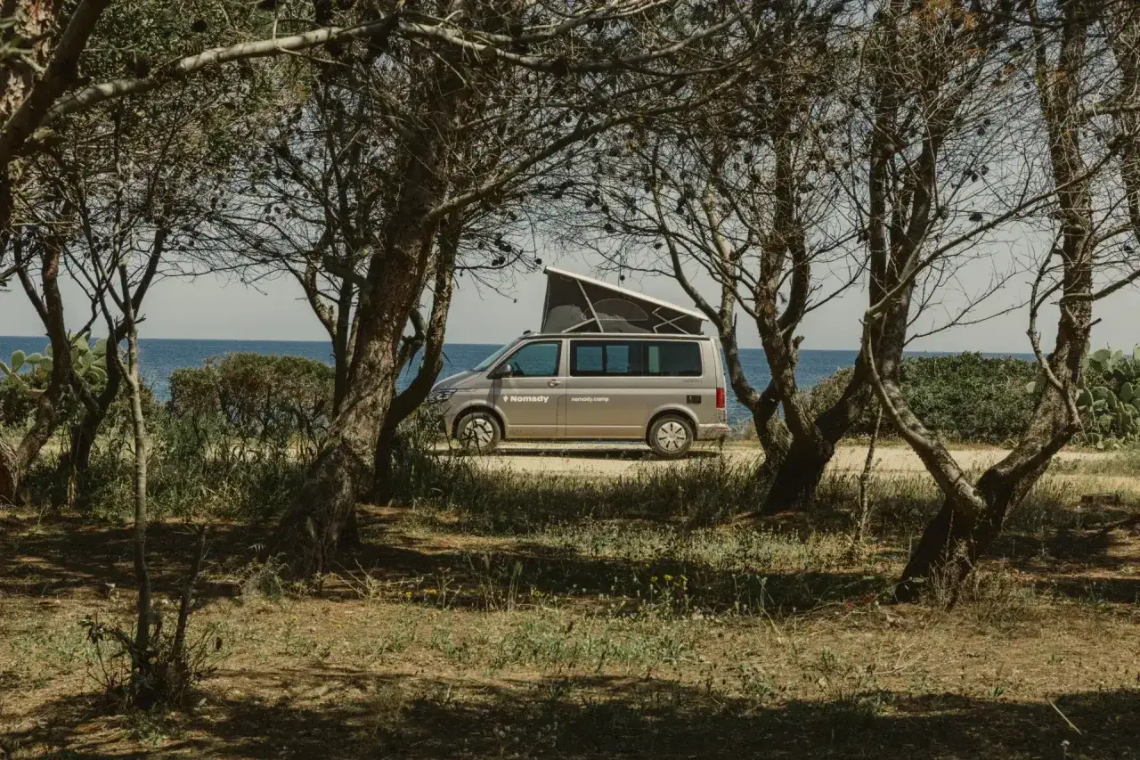 Ein Campervan steht unter B&auml;umen am Meer. Die mediterrane natur italien l&auml;dt zum Entspannen ein.