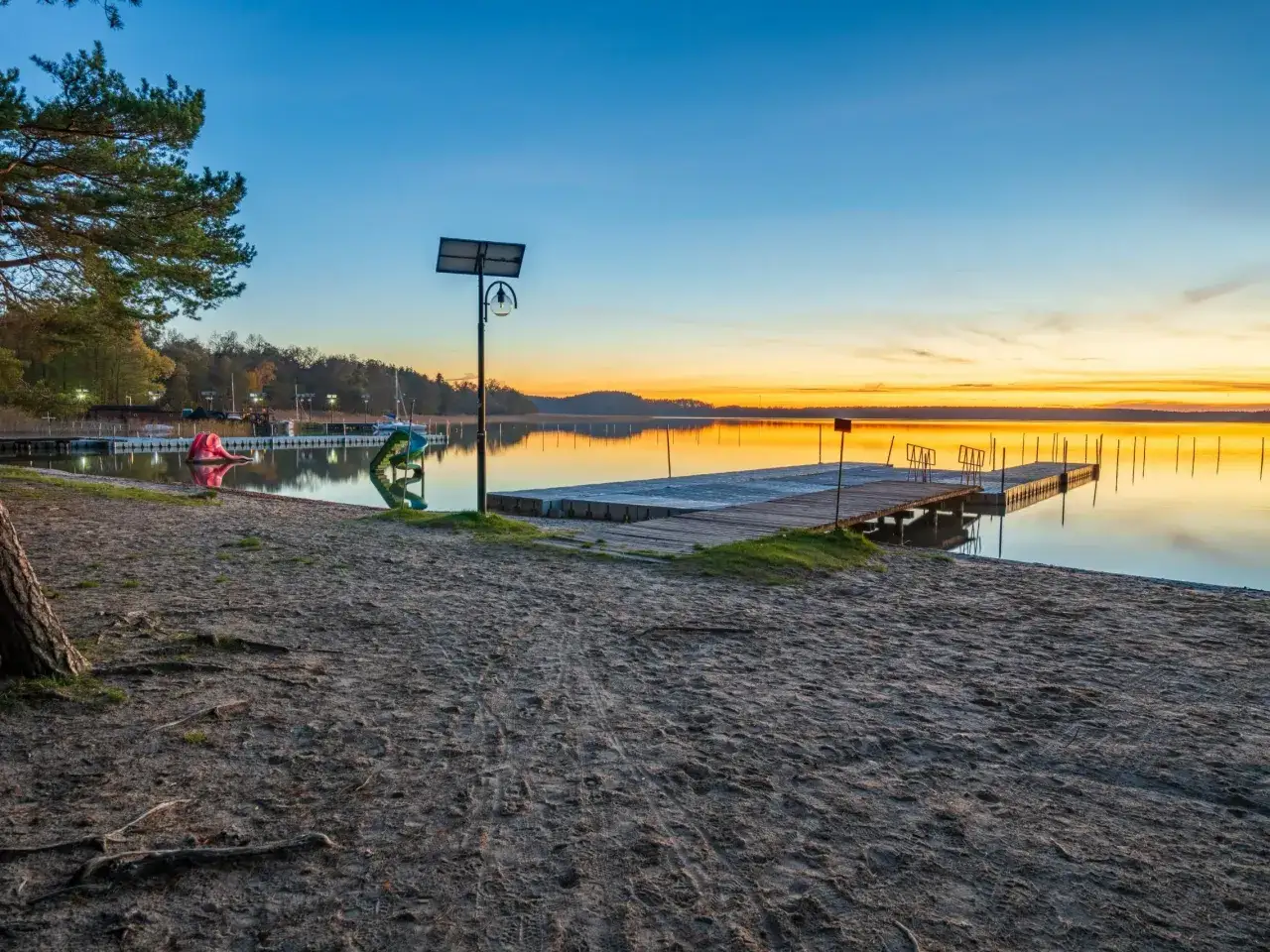 Piękny zachód słońca nad czystym jeziorem. Drewniane pomosty odbijają się w spokojnej tafli wody, a na brzegu widać piaszczystą plażę.