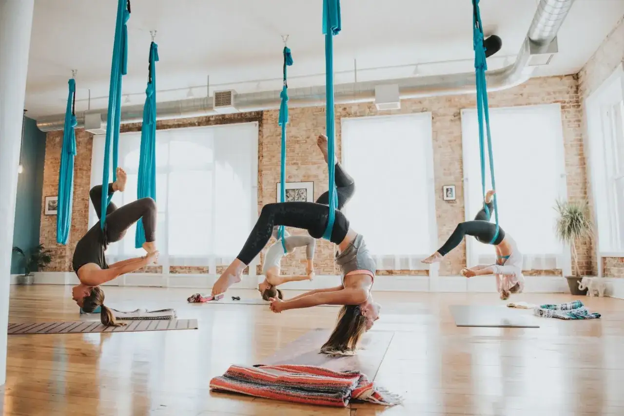 Kobiety ćwiczą **air yoga**, wisząc w błękitnych hamakach w przestronnej sali.