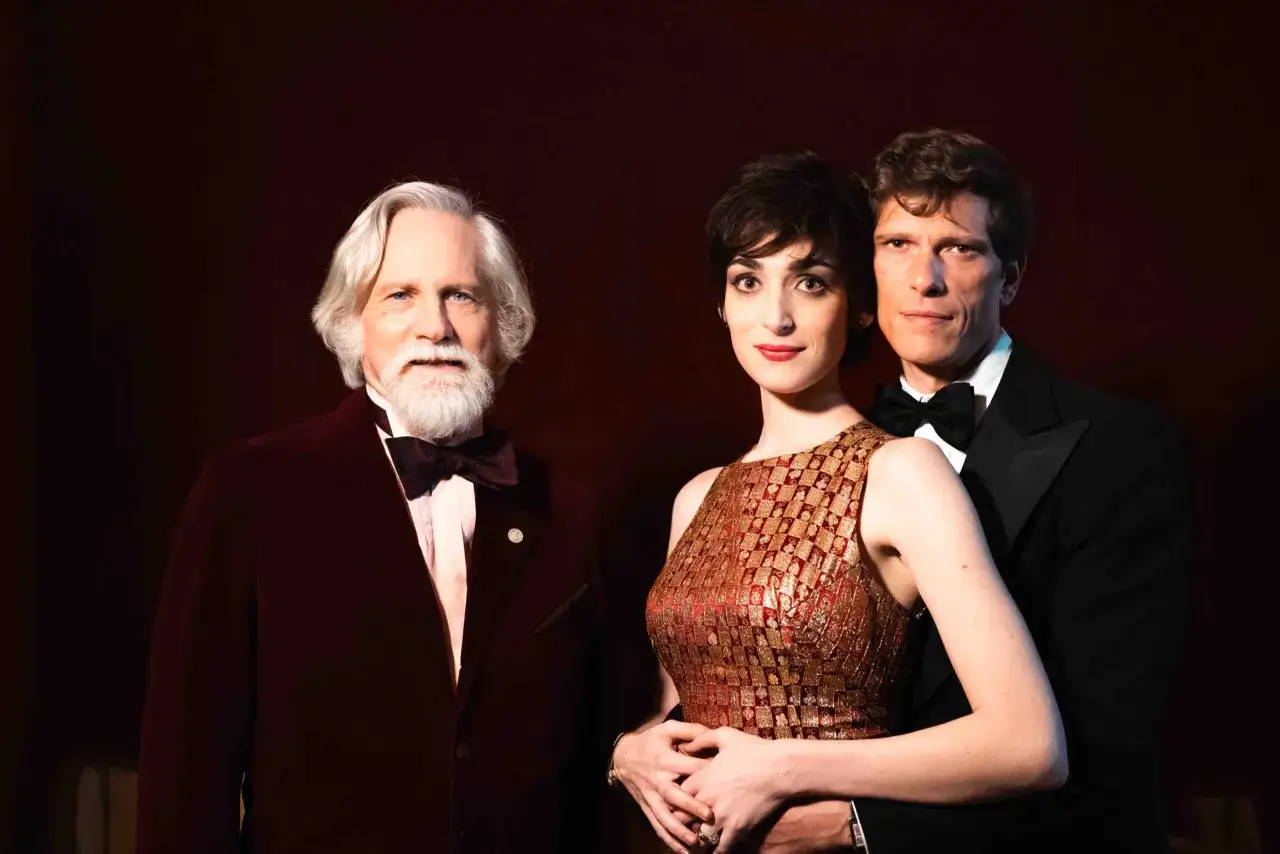 Tre figure eleganti, un uomo anziano con barba bianca e un completo bordeaux, una donna con abito dorato e un uomo con smoking. Anticipazioni cuori 3.