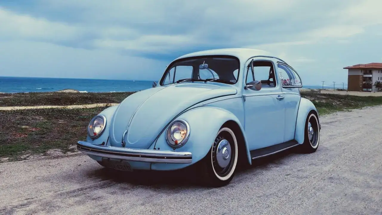 En ljusblå Volkswagen Bubbla, en ikonisk modell genom åren, parkerad vid havet.