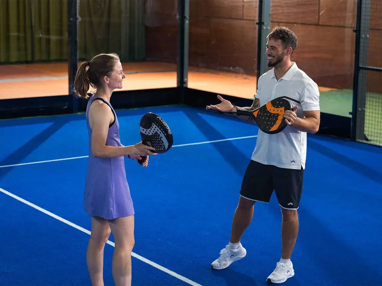 Coppia sorridente sul campo blu, pronta per una partita di padel. Lei con vestito viola, lui con polo bianca e pantaloncini neri.