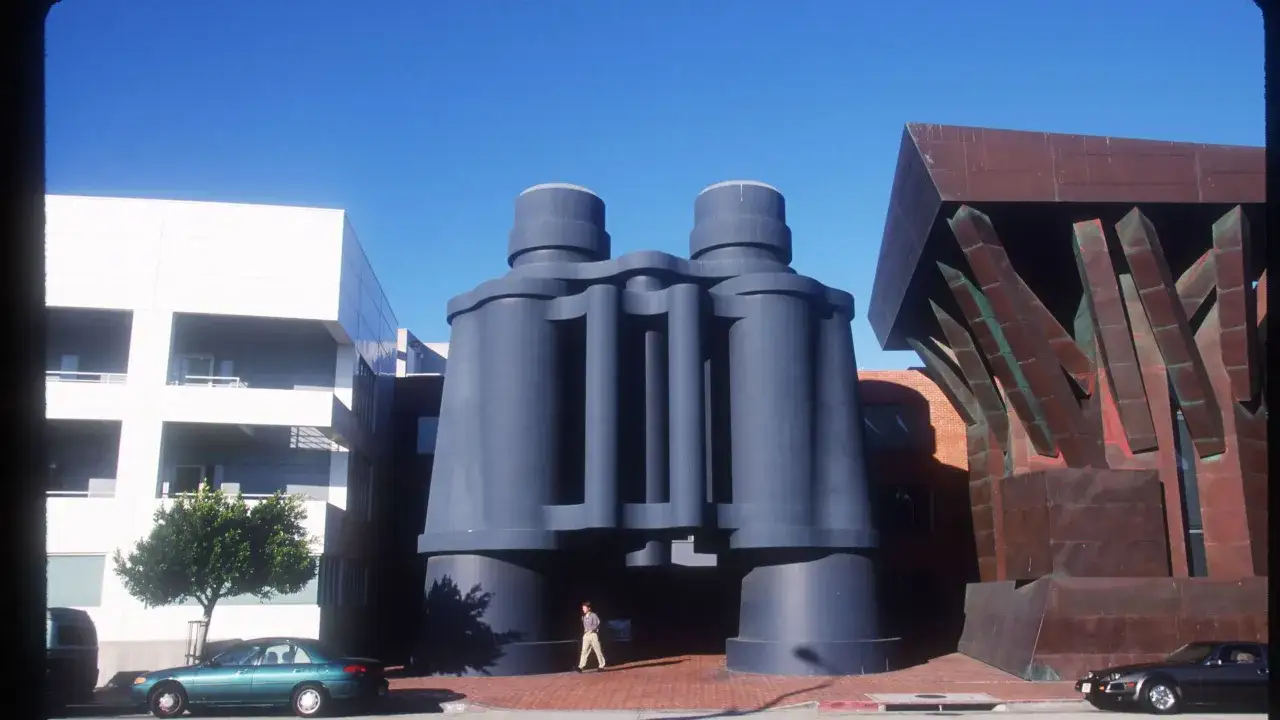 Monumentalne lornetka, symbol postmodernizmu, góruje nad ulicą. Obok nowoczesne budynki i rzeźbiarska fasada.