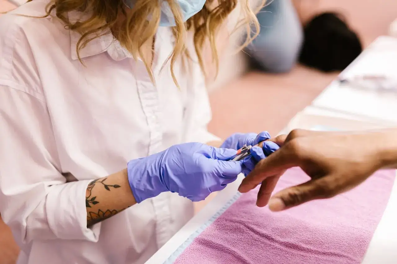 Manicurzystka w niebieskich rękawiczkach używa cążek do sk&oacute;rek, aby precyzyjnie usunąć nadmiar nask&oacute;rka wok&oacute;ł paznokcia klienta.